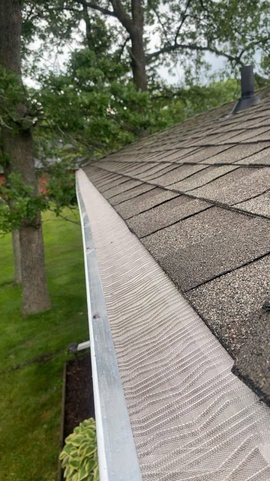 Close-up of a house roof with asphalt shingles and a white gutter beside trees and a grassy yard