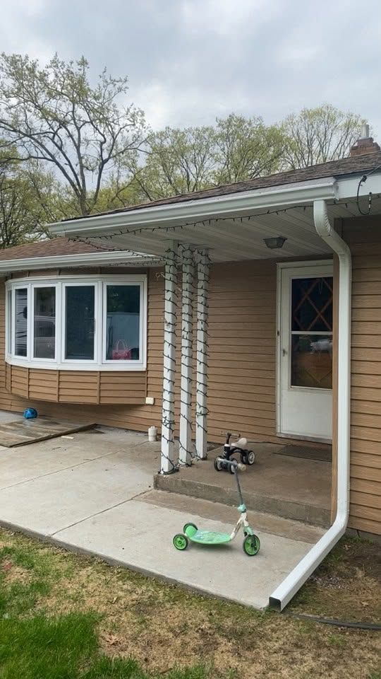 Front porch of a tan house with white-trimmed bay window, columns, and two scooters on the concrete step.