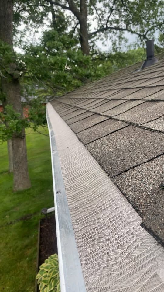 Rain gutter and shingled roof edge beside leafy trees and a green yard.