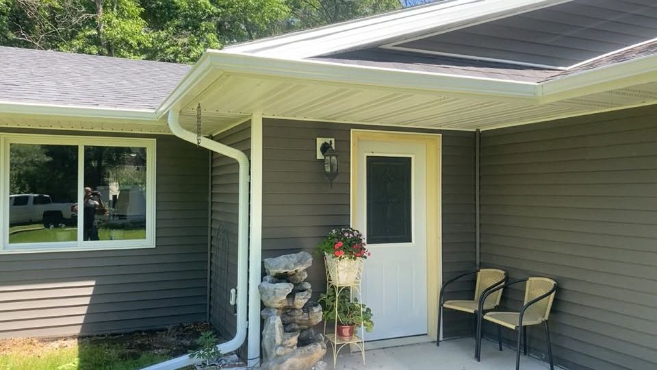 Front porch with white door, hanging flowers, two chairs, and a large window on a gray house.