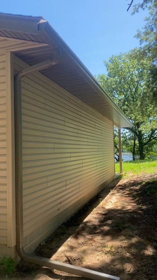 Side of a beige house with white trim beside a dirt path and green trees under a blue sky