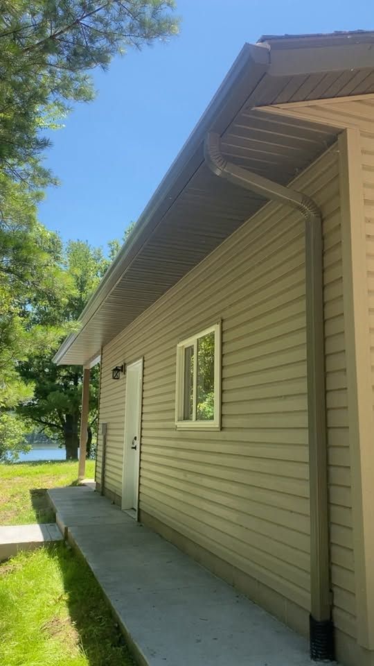 Side of a beige house with vinyl siding, white-trim windows, gutter, and a concrete walkway beside green grass and trees.