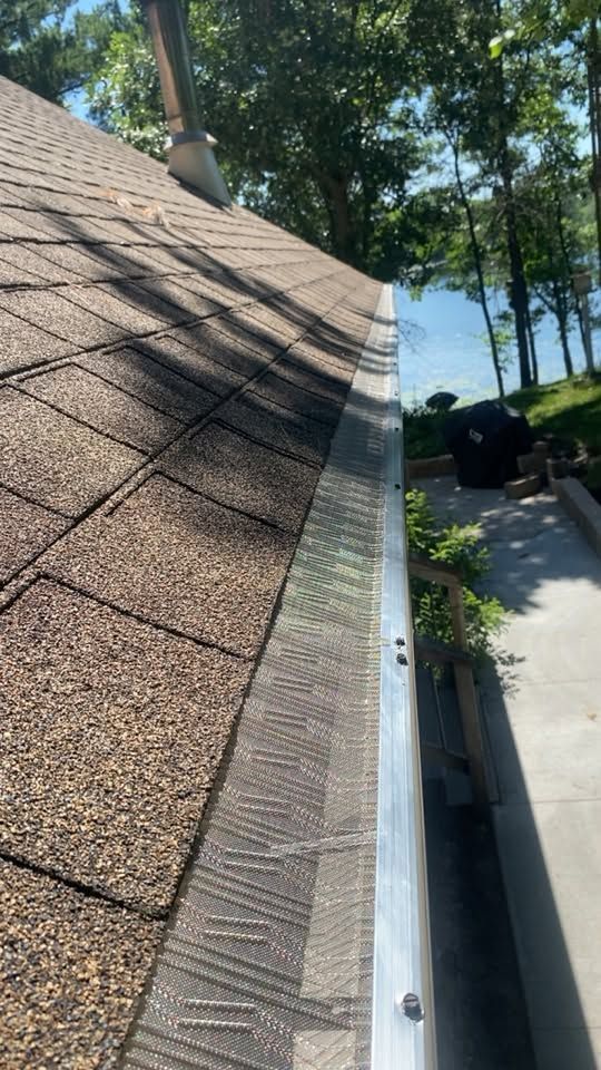 Close view of a shingled roof edge with gutter, overlooking a lakeside yard and trees.