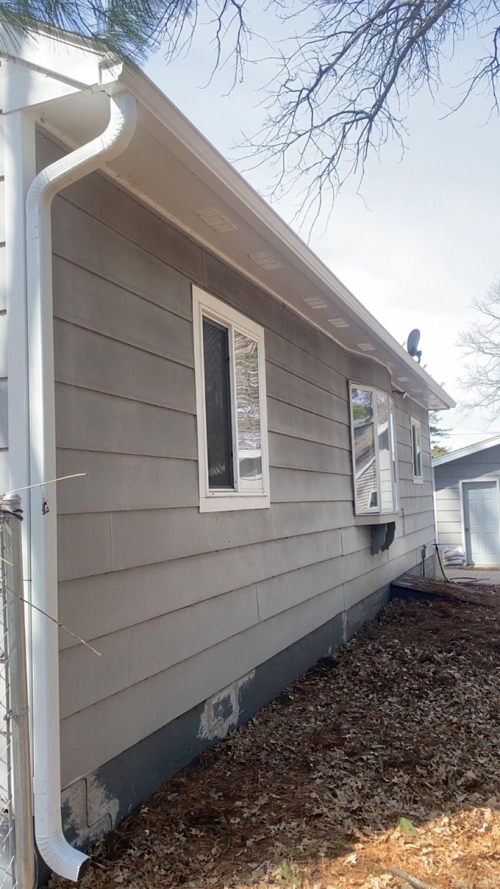 Side view of a gray house with white trim, windows, gutter, and leaf-covered ground beside it