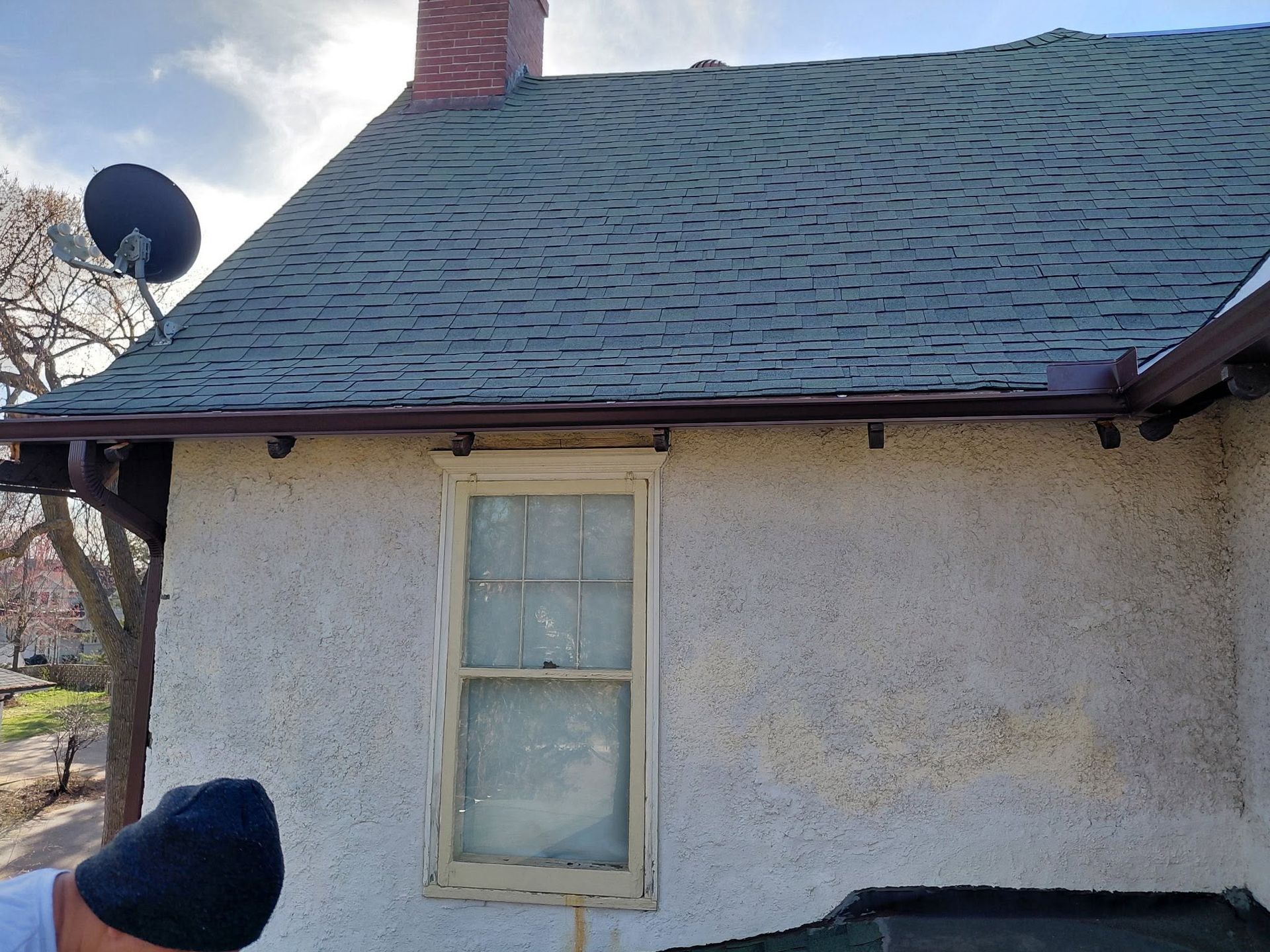 Exterior wall of a house with a small window, shingled roof, chimney, and satellite dish in daylight