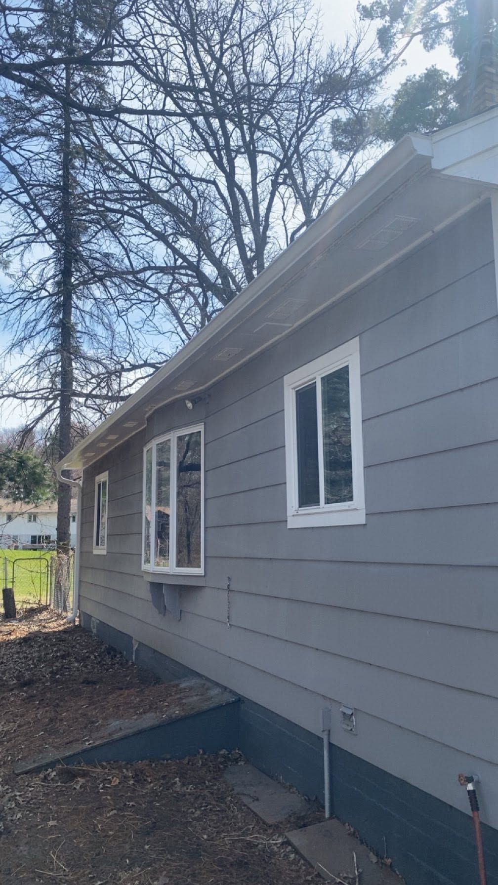 Gray house exterior with white-trimmed windows, blue trim, and a leaf-covered yard along the side.