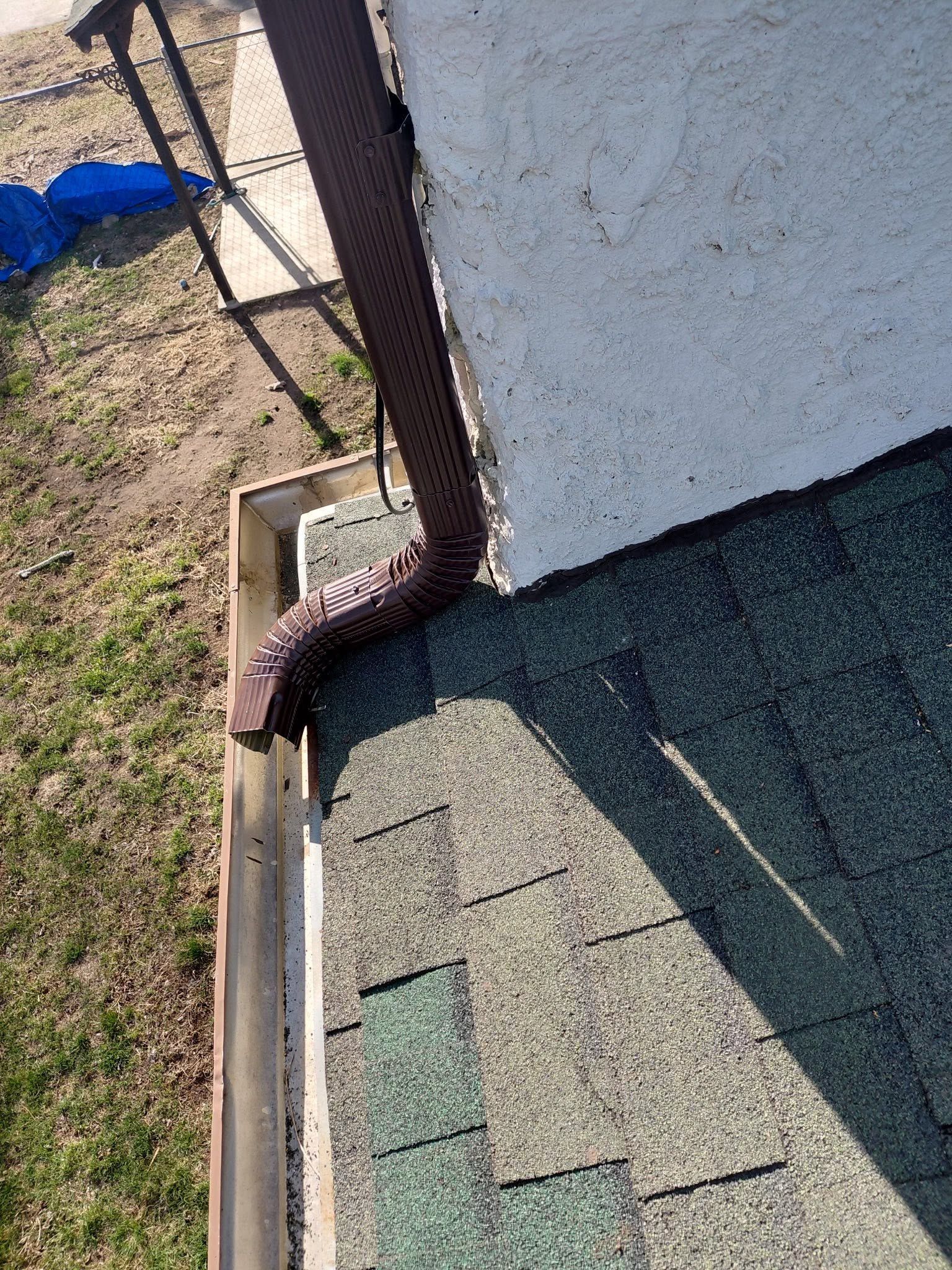 Roof gutter and downspout beside a white wall, draining toward a grassy yard with a blue tarp nearby.