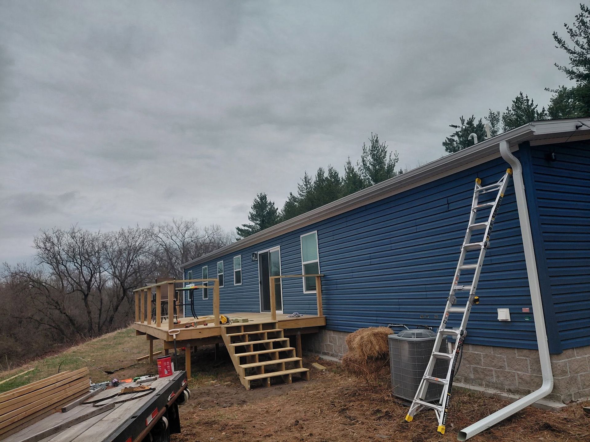 Blue house exterior with a new wooden deck and stairs, plus ladders and a downspout along the side.