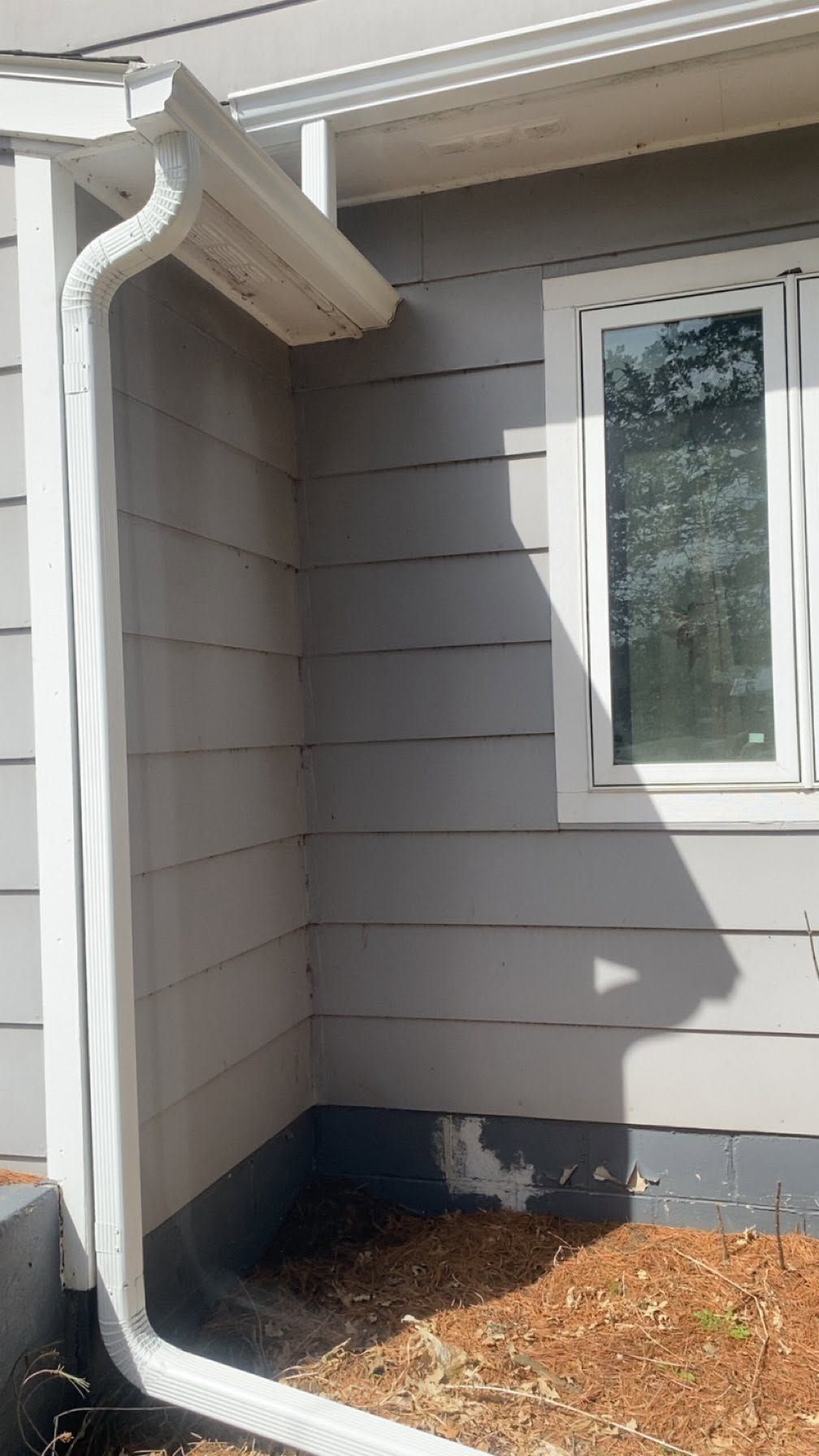 Exterior house wall with white downspout, window, gray siding, and mulch bed at the foundation