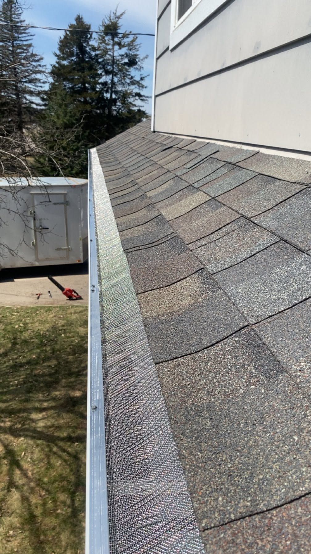 Row of gray shingle roof along a white building wall, with trees and blue sky in the background.