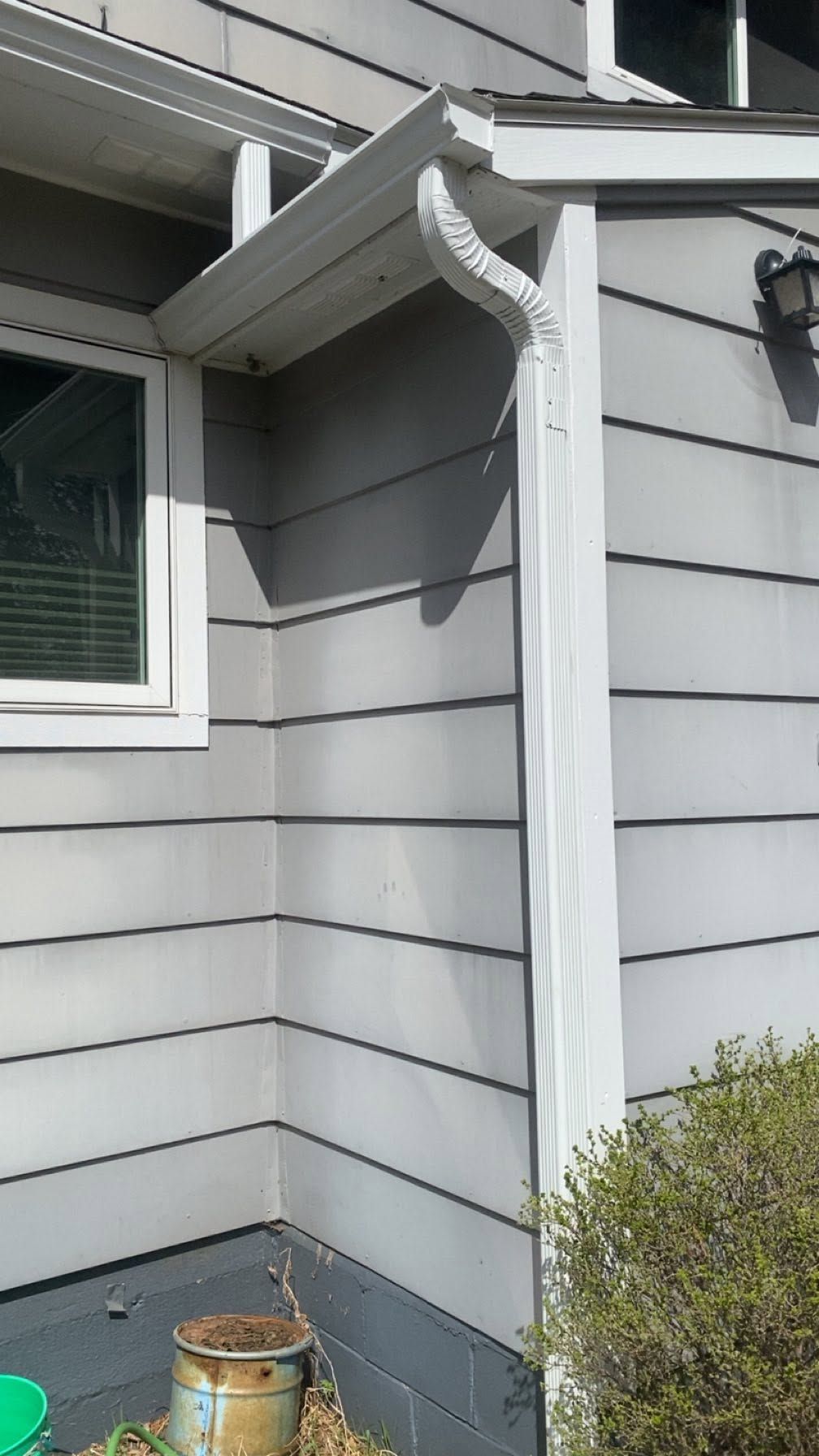 Damaged white gutter and downspout on a gray house exterior, with a bucket and shrub below