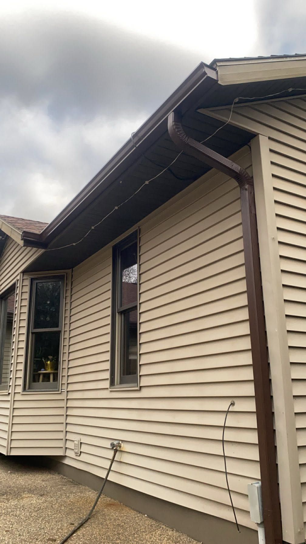 Tan house siding with windows, gutter, and downspout under a cloudy sky