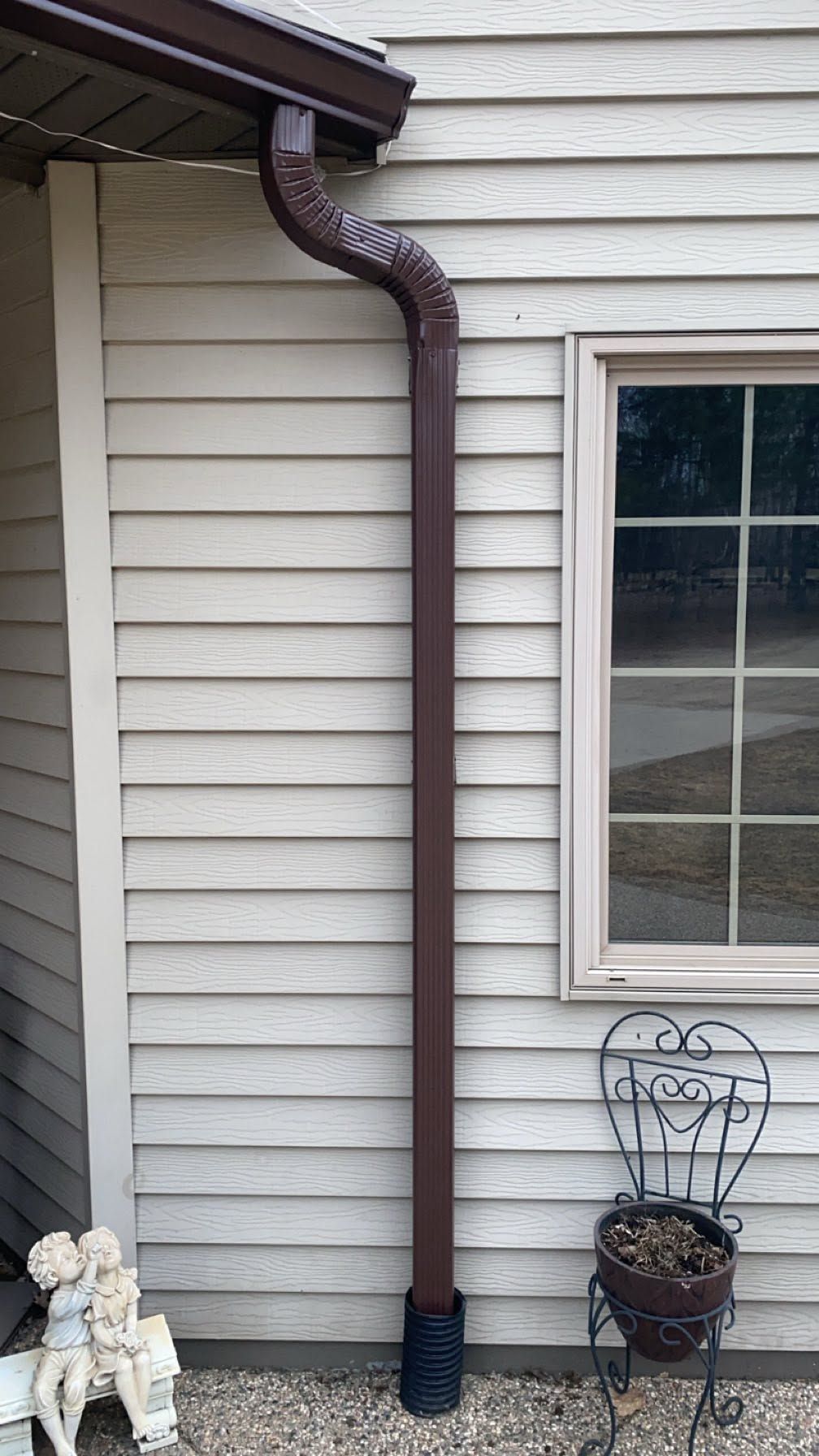 Brown downspout with elbow attached to beige house siding beside a window and decorative planter basket
