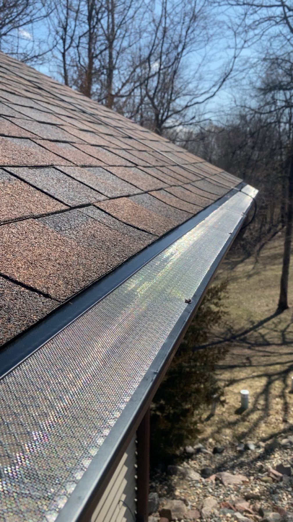 Close-up of a roof edge with metal gutter and leaf guard, bordered by brown shingles and winter trees below