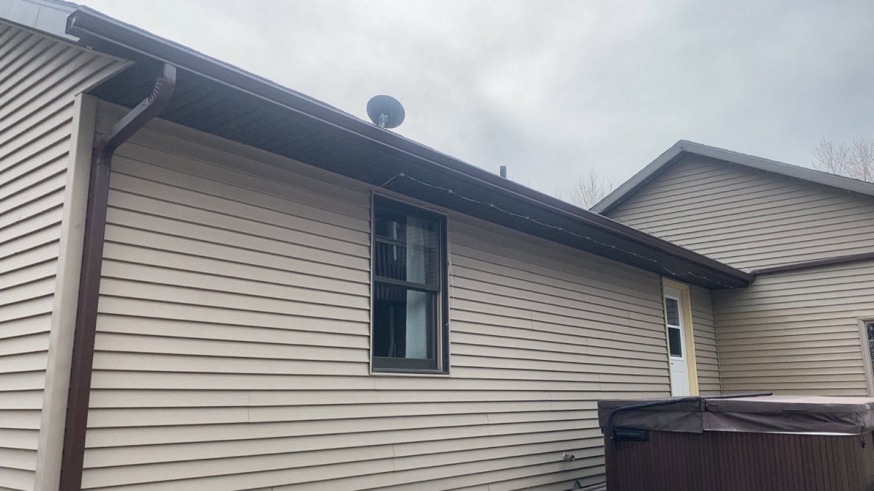 Side view of a beige house with vinyl siding, a narrow window, roof vent, and overcast sky.