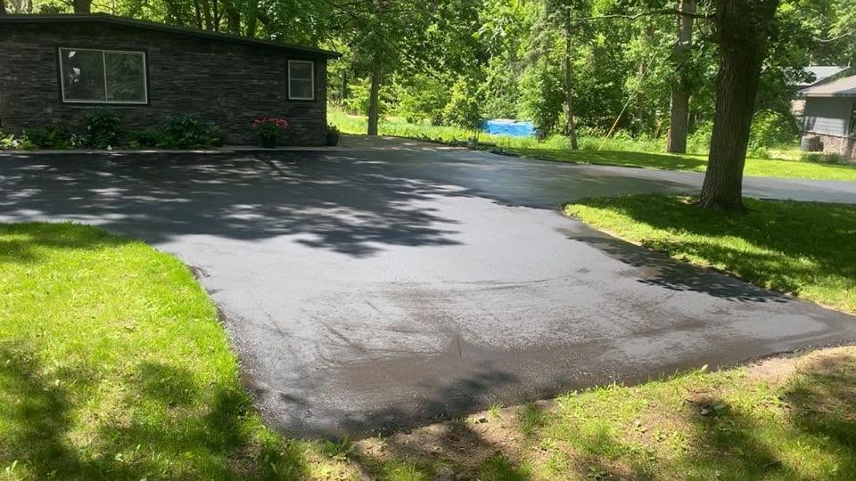 Freshly paved driveway beside a dark house in a sunny, tree-lined yard