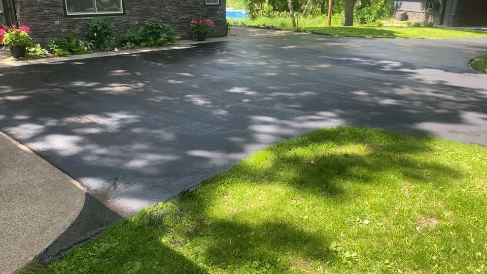 Quiet suburban driveway beside a green lawn, with a house and tree shadows in sunlight.