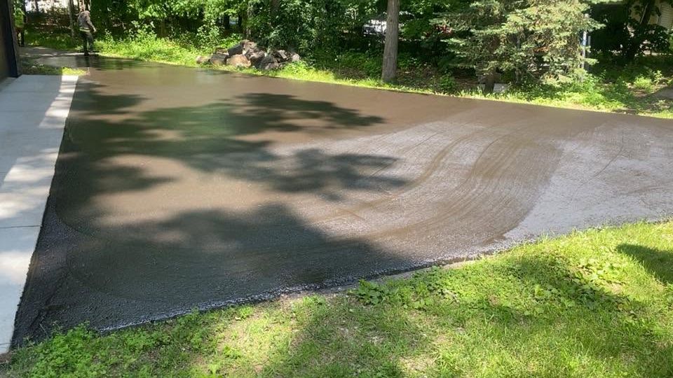 Freshly paved driveway or patio with wet, dark asphalt beside a green lawn and trees