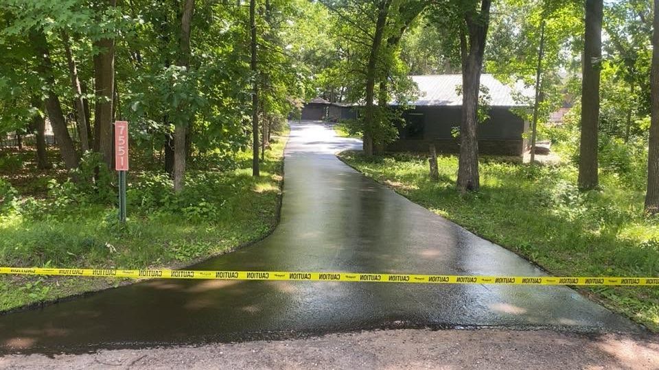 Yellow caution tape blocks a wet driveway in a wooded area, with a small building in the background.
