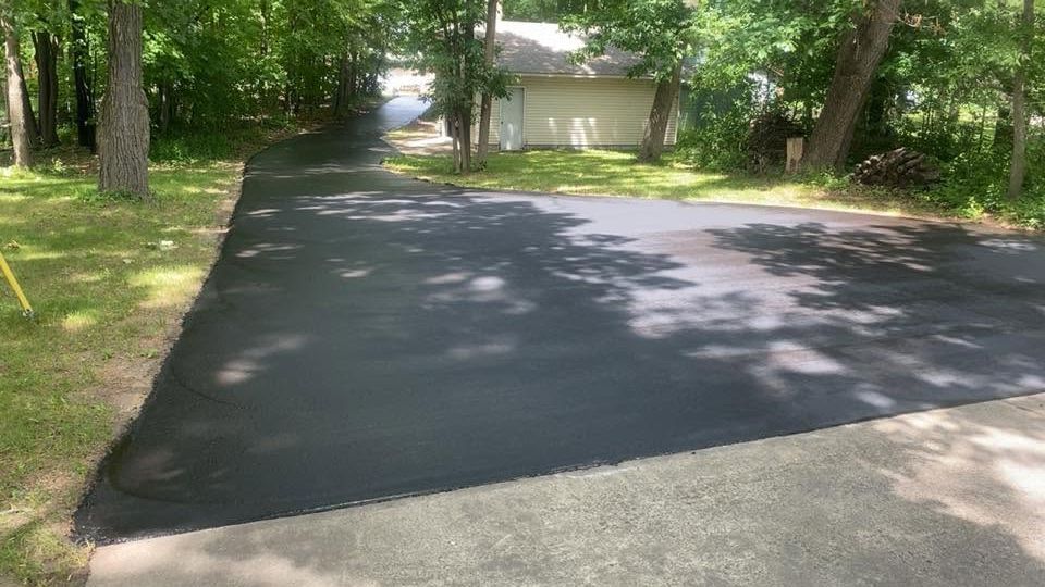 Freshly paved driveway in a wooded residential area, with a small building in the background.