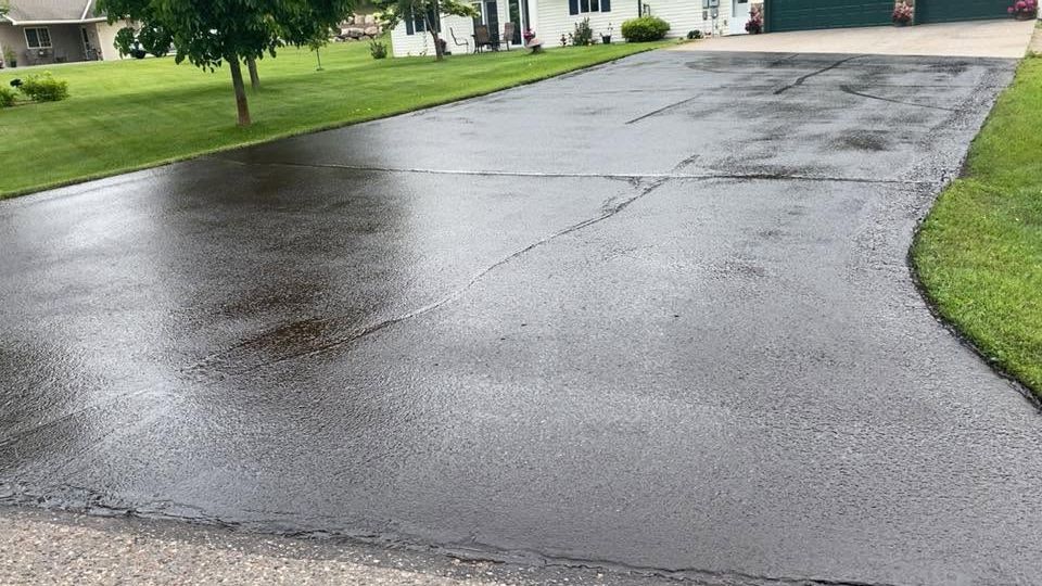 Wet black asphalt driveway after rain, with green lawns and houses along the sides