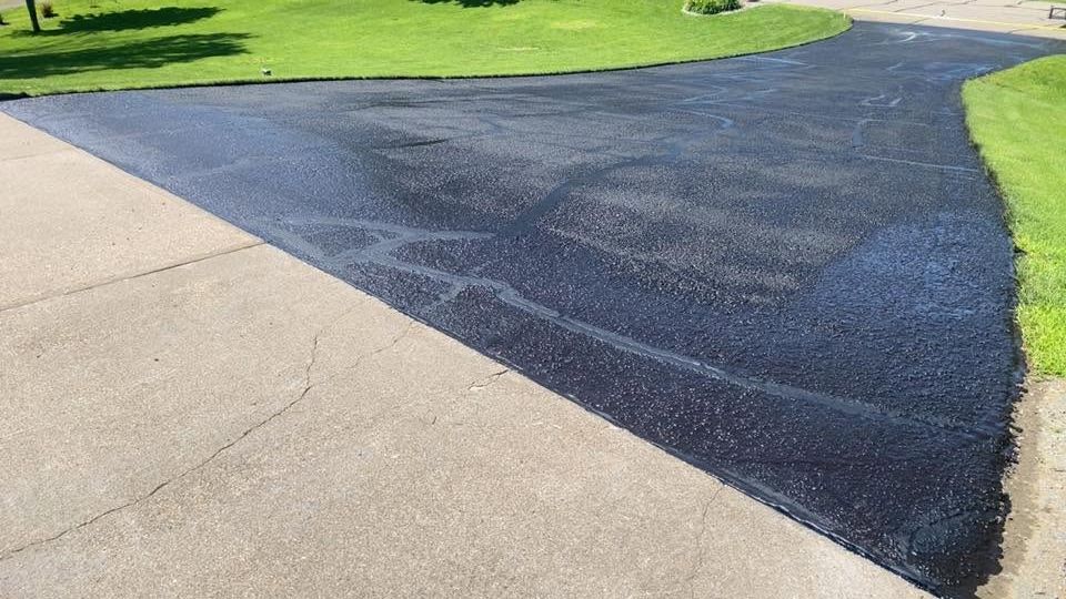 Freshly sealed dark asphalt patch meeting a light concrete driveway beside green lawn