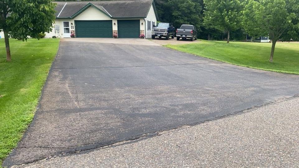 Wide asphalt driveway leading to a two-car garage with two parked vehicles and green trees around it.