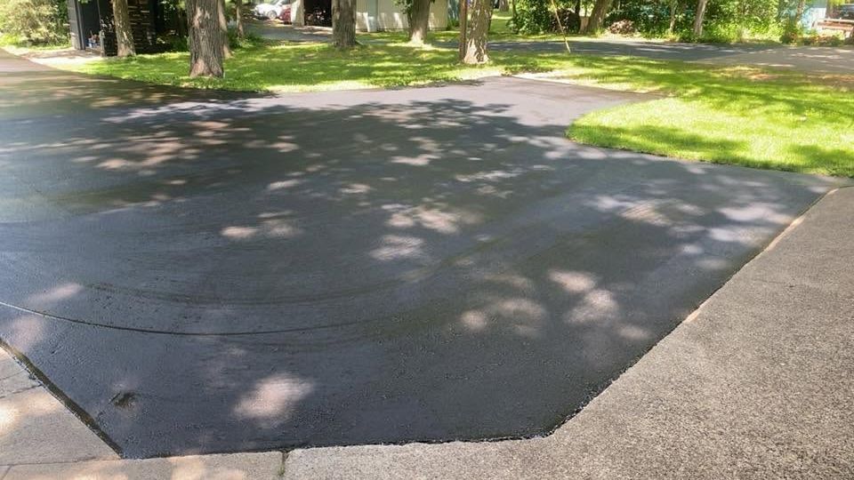 Fresh black asphalt driveway bordered by concrete, with trees casting dappled shadows in a park-like setting