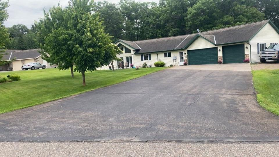 Suburban house with a long asphalt driveway, green lawn, and trees under cloudy skies