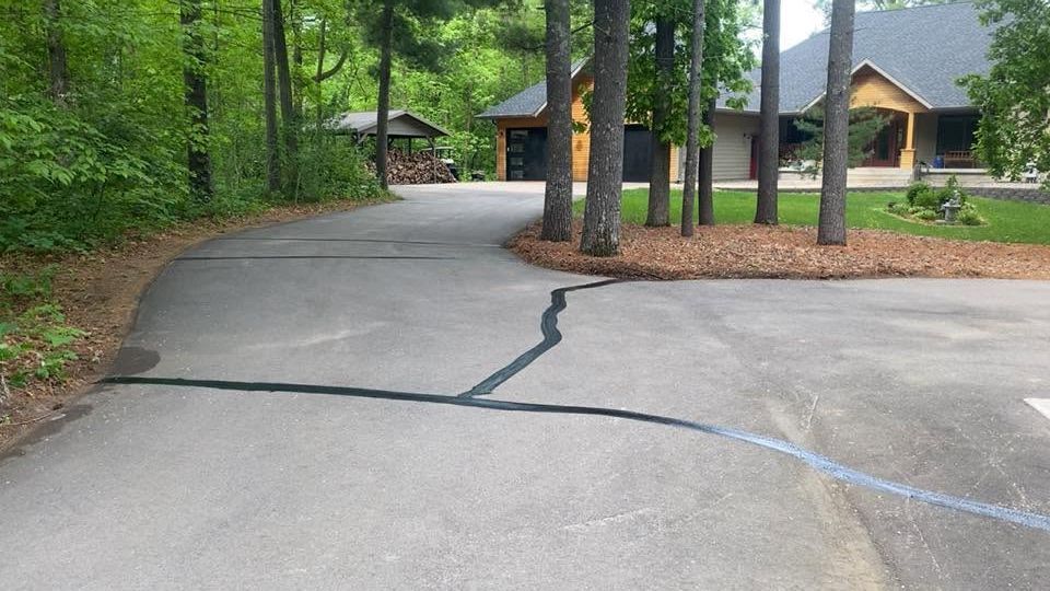 Cracked concrete driveway curving toward a lodge-style house in a wooded setting