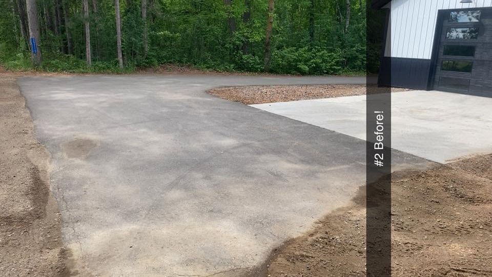 Freshly poured concrete driveway beside a white building, bordered by dirt and trees