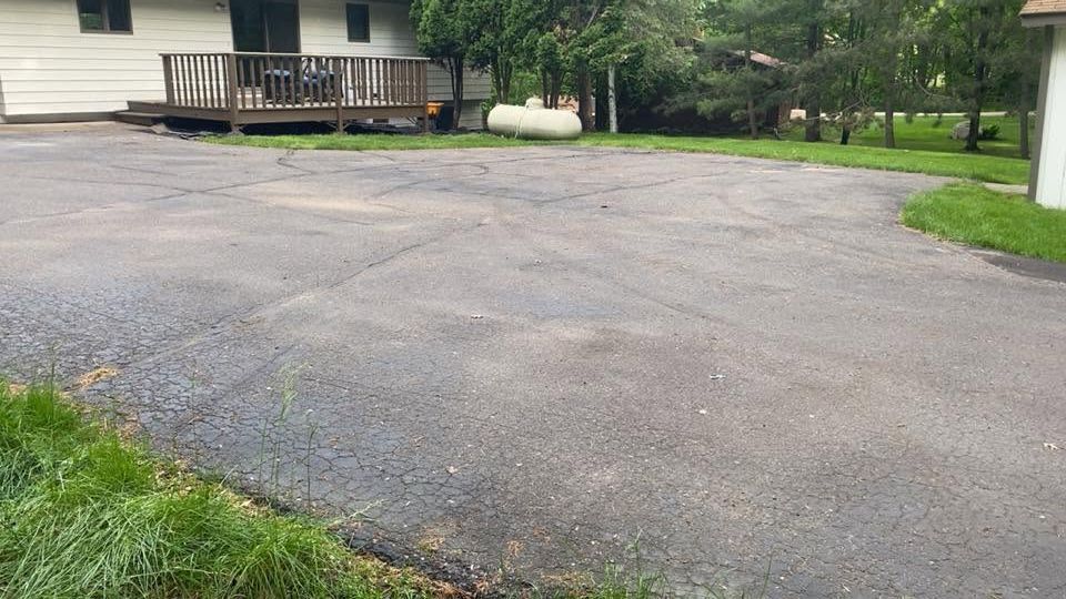 Empty asphalt driveway beside a house with a wooden deck and surrounding grass and trees
