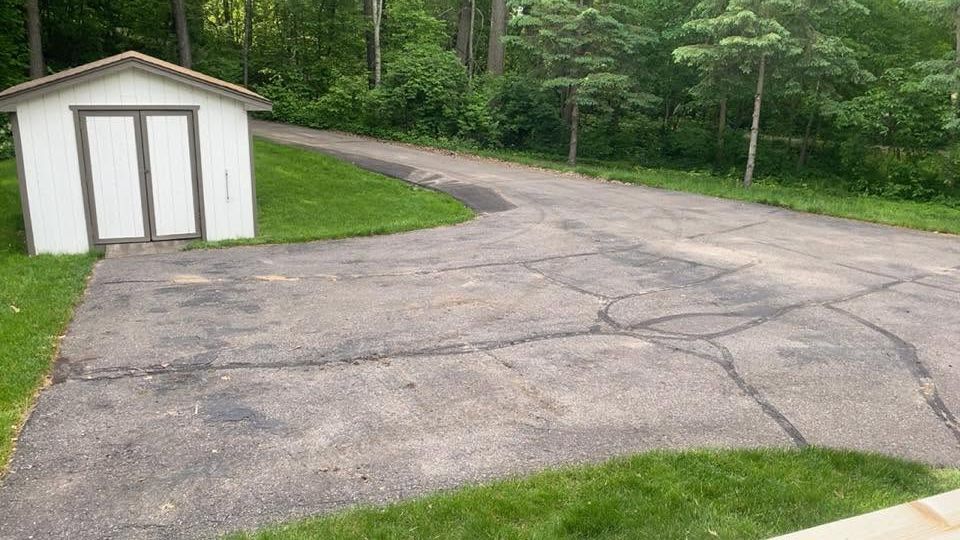 Cracked asphalt driveway beside a small white shed, curving into a wooded area.