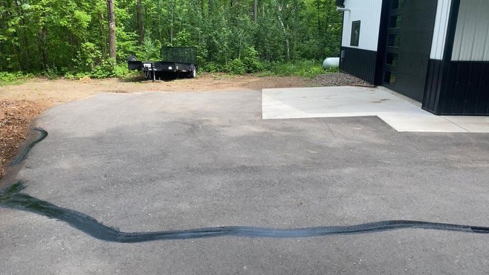 Driveway beside a white-and-black building, bordered by trees and a black hose or cable across the pavement.
