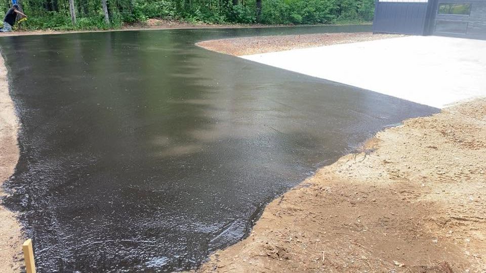 Freshly paved black surface beside a sandy bank and white concrete area near a wooded shoreline