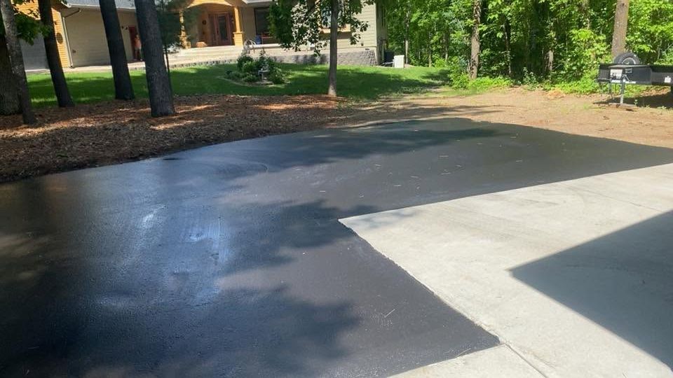 Freshly paved driveway beside a concrete sidewalk, with trees and a house in the background