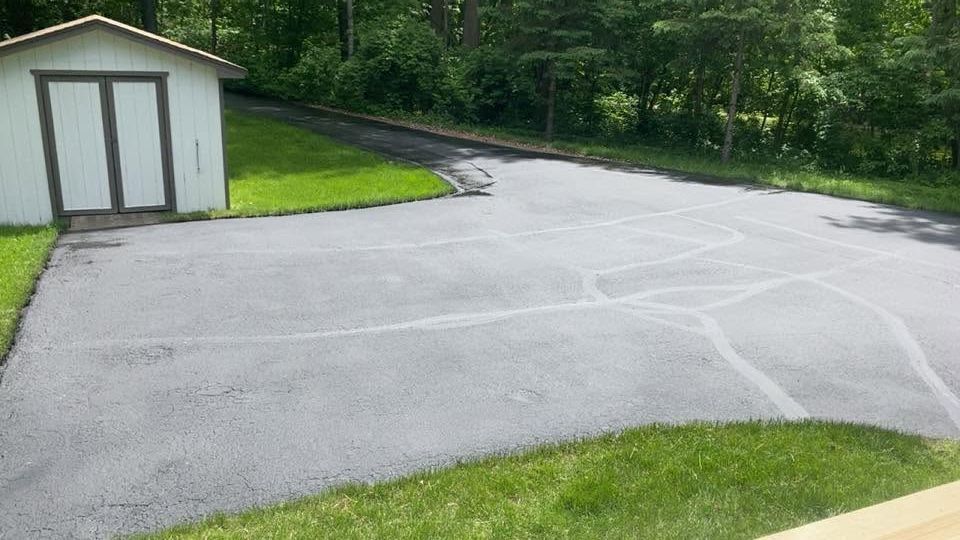 Empty paved driveway beside a small shed, bordered by grass and trees.