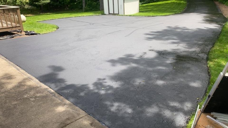 Freshly paved black driveway with tree shadows, grass border, and a concrete walkway beside it
