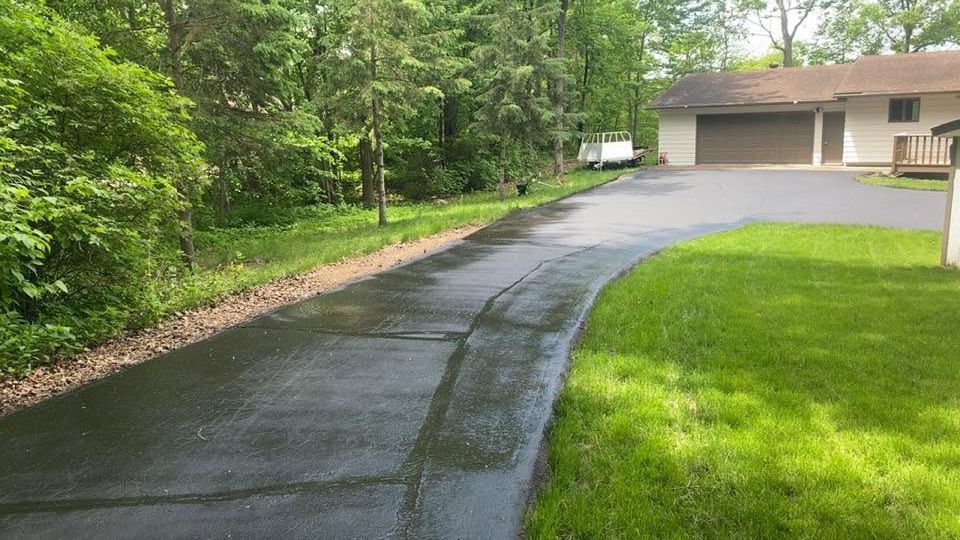Freshly paved black driveway curving past green lawn and trees beside a suburban house