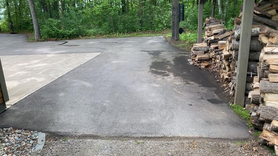 Freshly paved asphalt driveway beside stacked firewood and trees