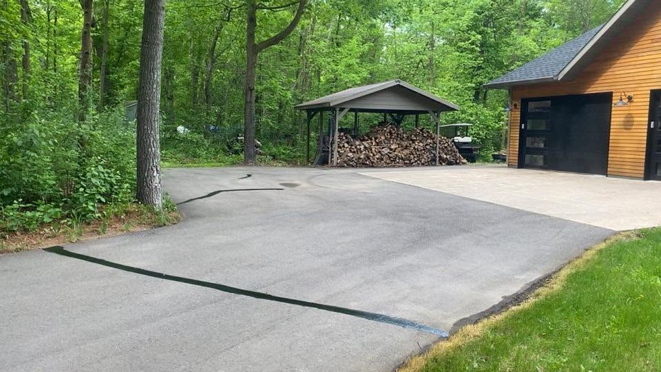 Driveway beside a garage with a woodpile shelter, surrounded by green trees and grass