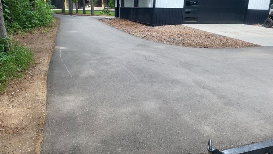 Empty asphalt driveway beside a house, with gravel and greenery along the edges.