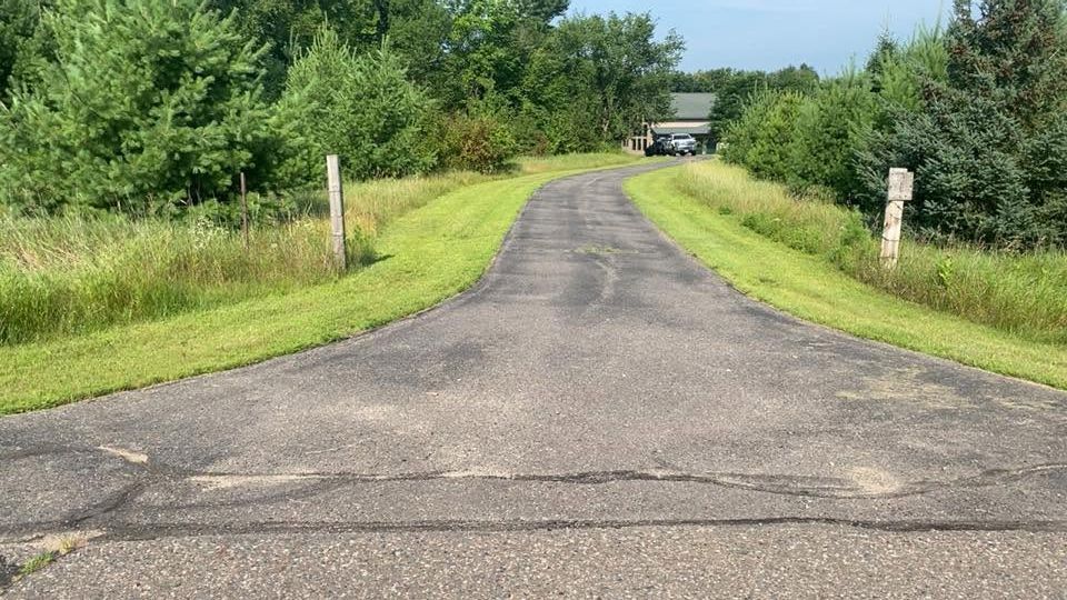 Narrow paved road curving through green grass and trees toward a small house in the distance