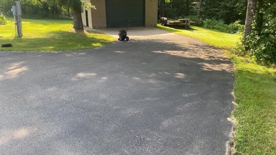 Black dog sitting on a driveway in front of a garage, with grass and trees nearby.