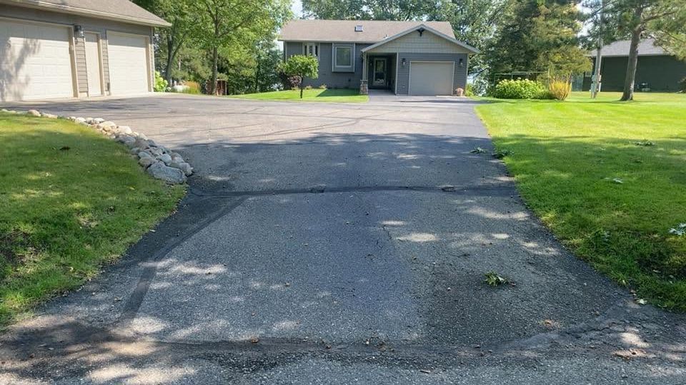 Cracked asphalt driveway leading to a garage, with green lawns and houses on both sides.