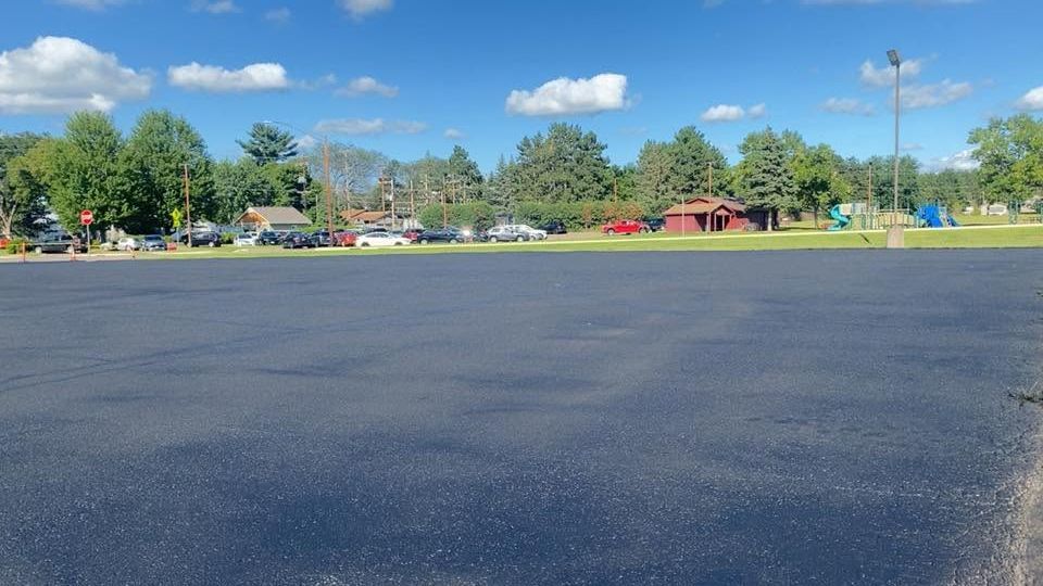 Empty paved lot beside trees and red buildings under a bright blue sky with clouds