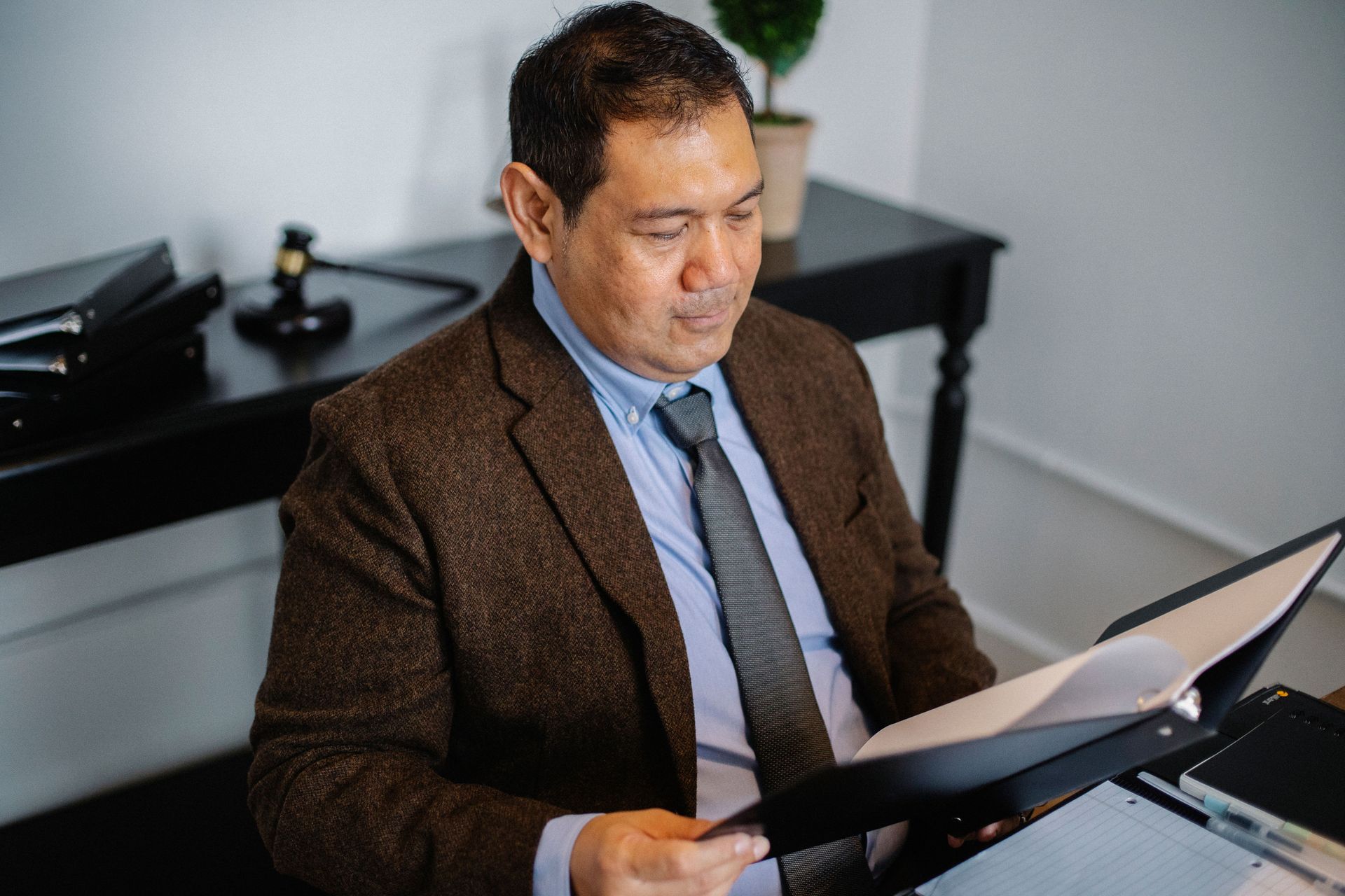 Man in a tweed jacket and tie seated at a desk, reading through a document in an open ring binder
