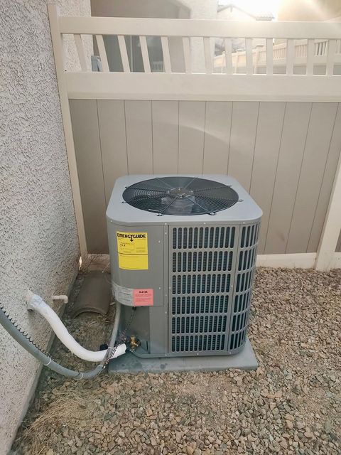 A gray outdoor air conditioning unit sits on a gravel surface against a wall and a beige fence.