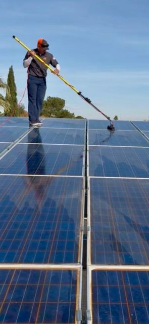 A person in a gray shirt and hat uses a long-handled cleaning brush to scrub a large array of rooftop solar panels.