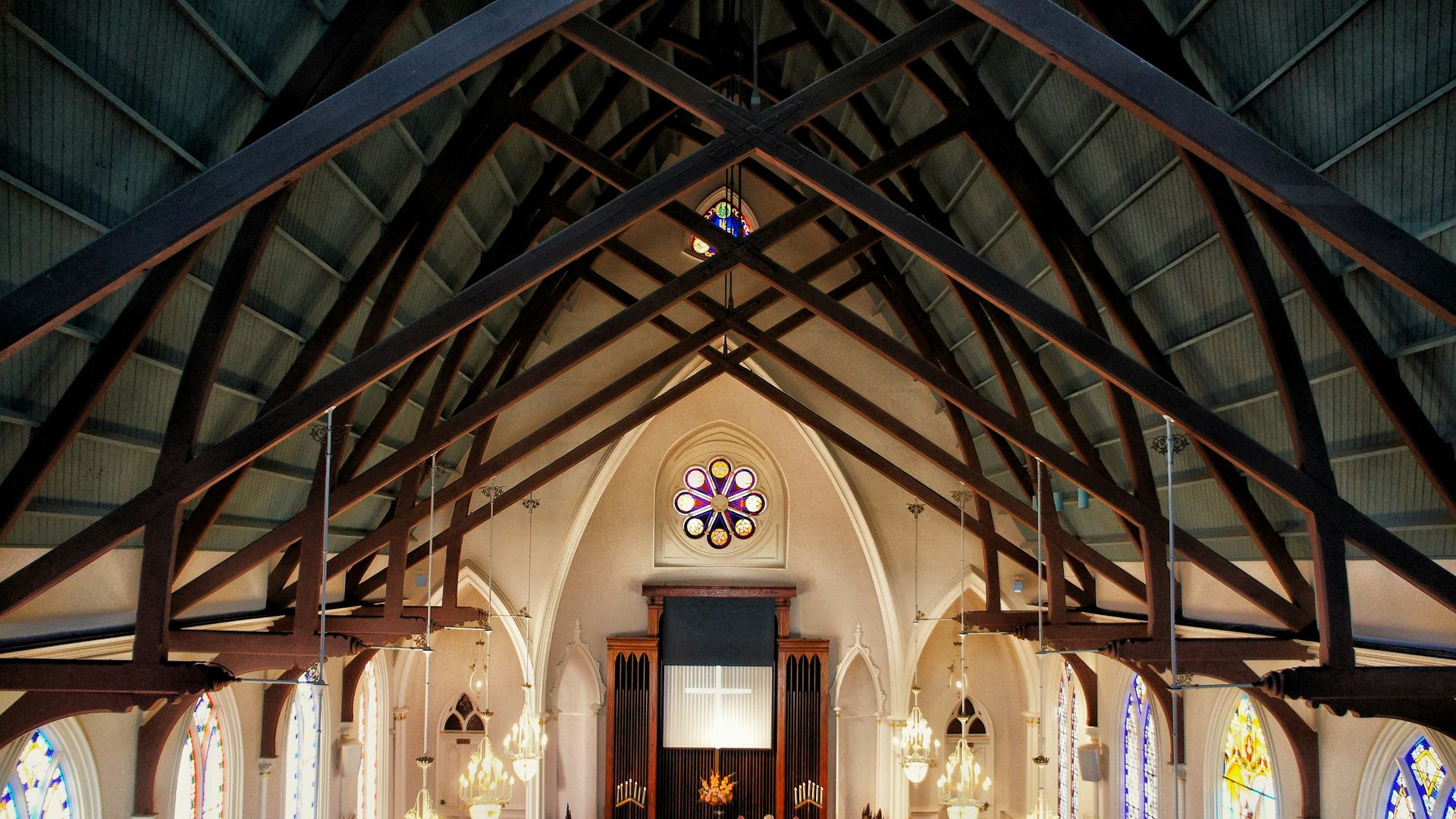 Wooden church ceiling with intricate beams, stained-glass window, and ornate chandeliers.